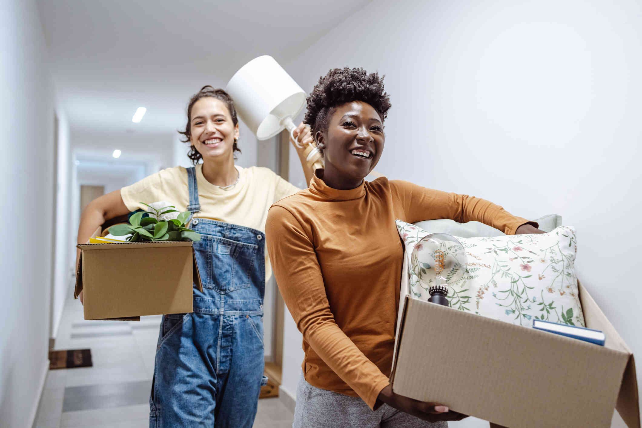 A girl in overalls holds a cardboard box with plants in it and a lamp. Another girl in an orange shirt holds a cardboard box with a pillow in it and stands in the hallway in front of the other girl smiling.